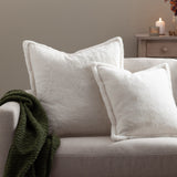 Two Kallu reversible faux fur cushions in ecru with Oxford flange borders, styled on a beige sofa beside a green throw and lit side table.