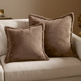 Two square Kallu faux fur cushions in mocha brown on a beige sofa, showing soft pile texture in two sizes with matching finish on both sides.