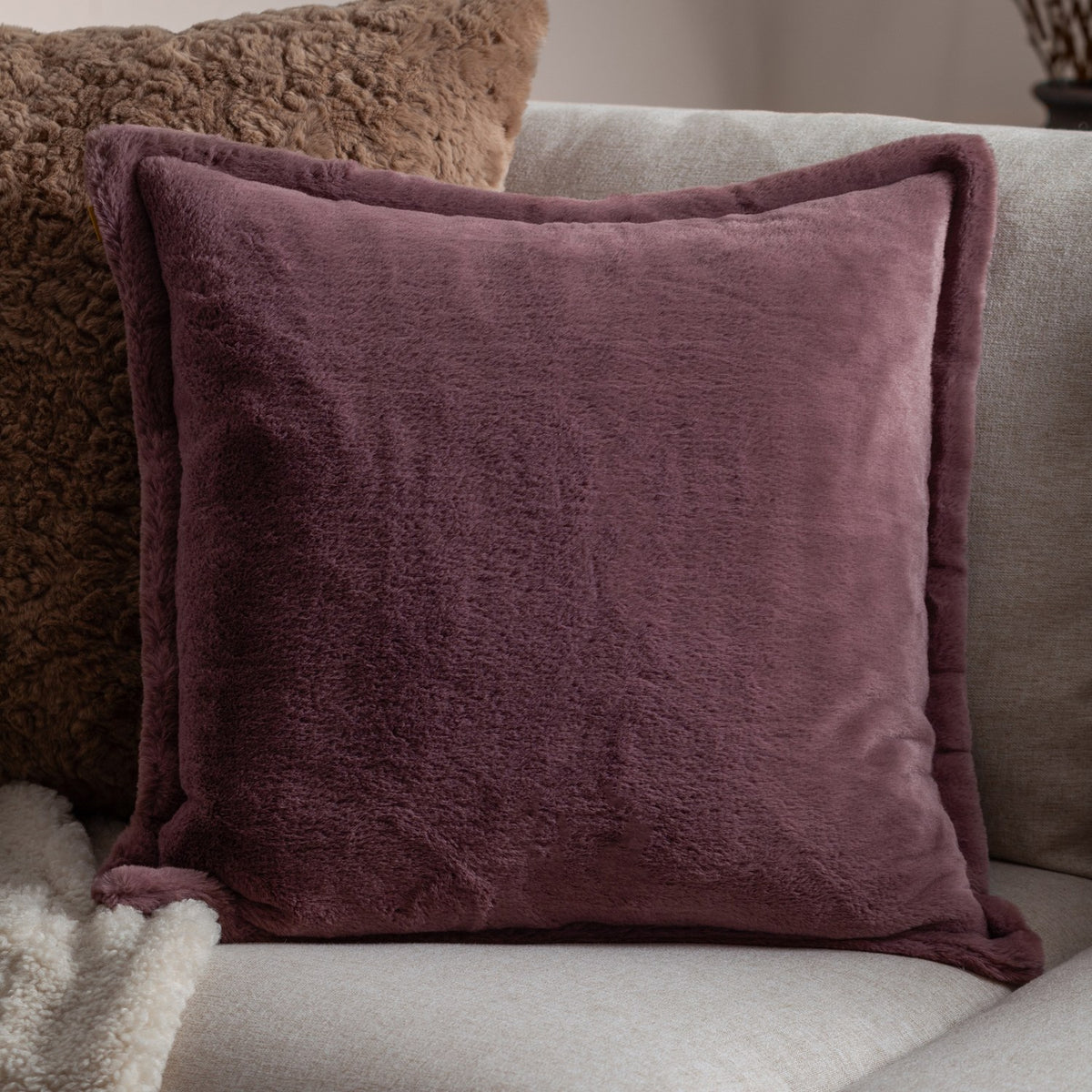 Kallu Reversible Faux Fur Cushion in mulberry with an Oxford flange border, shown on a light grey sofa alongside a brown textured cushion and cream throw.