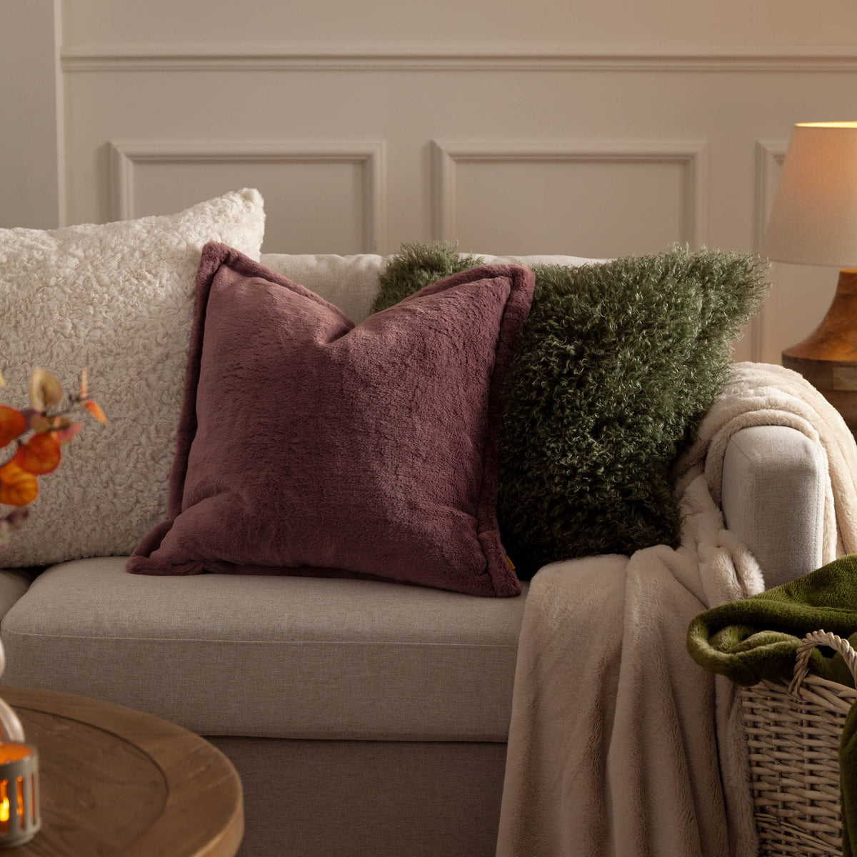 Kallu Reversible Faux Fur Cushion in mulberry with an Oxford flange border, styled on a beige sofa alongside cream and green textured cushions.