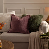 Kallu Reversible Faux Fur Cushion in mulberry with an Oxford flange border, styled on a beige sofa alongside cream and green textured cushions.