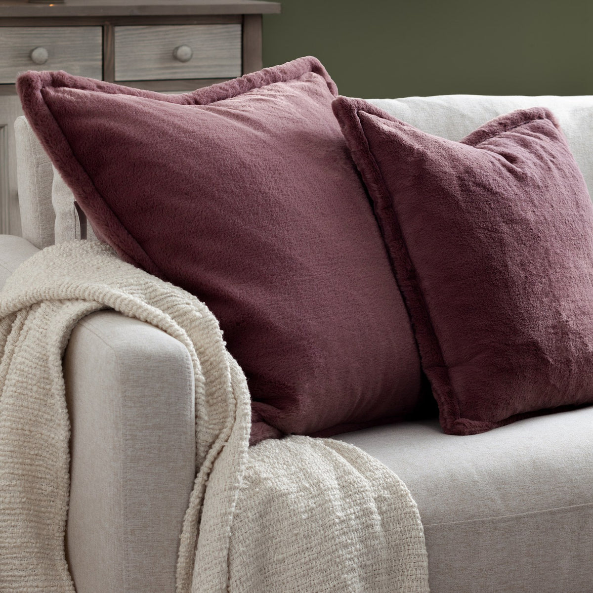 Kallu Reversible Faux Fur Cushion in mulberry with Oxford flange border, styled on a light grey sofa with a cream throw in a living room setting.