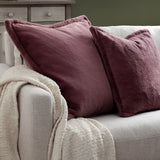Kallu Reversible Faux Fur Cushion in mulberry with Oxford flange border, styled on a light grey sofa with a cream throw in a living room setting.