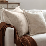 Two Kallu reversible faux fur cushions in natural beige with plush pile and Oxford flange edging, styled on a cream sofa with a brown throw.
