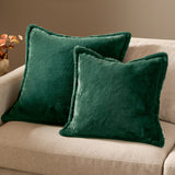 Two square Kallu faux fur cushions in needle green with plush pile and raised border edging, styled on a beige sofa beside a dried flower arrangement.