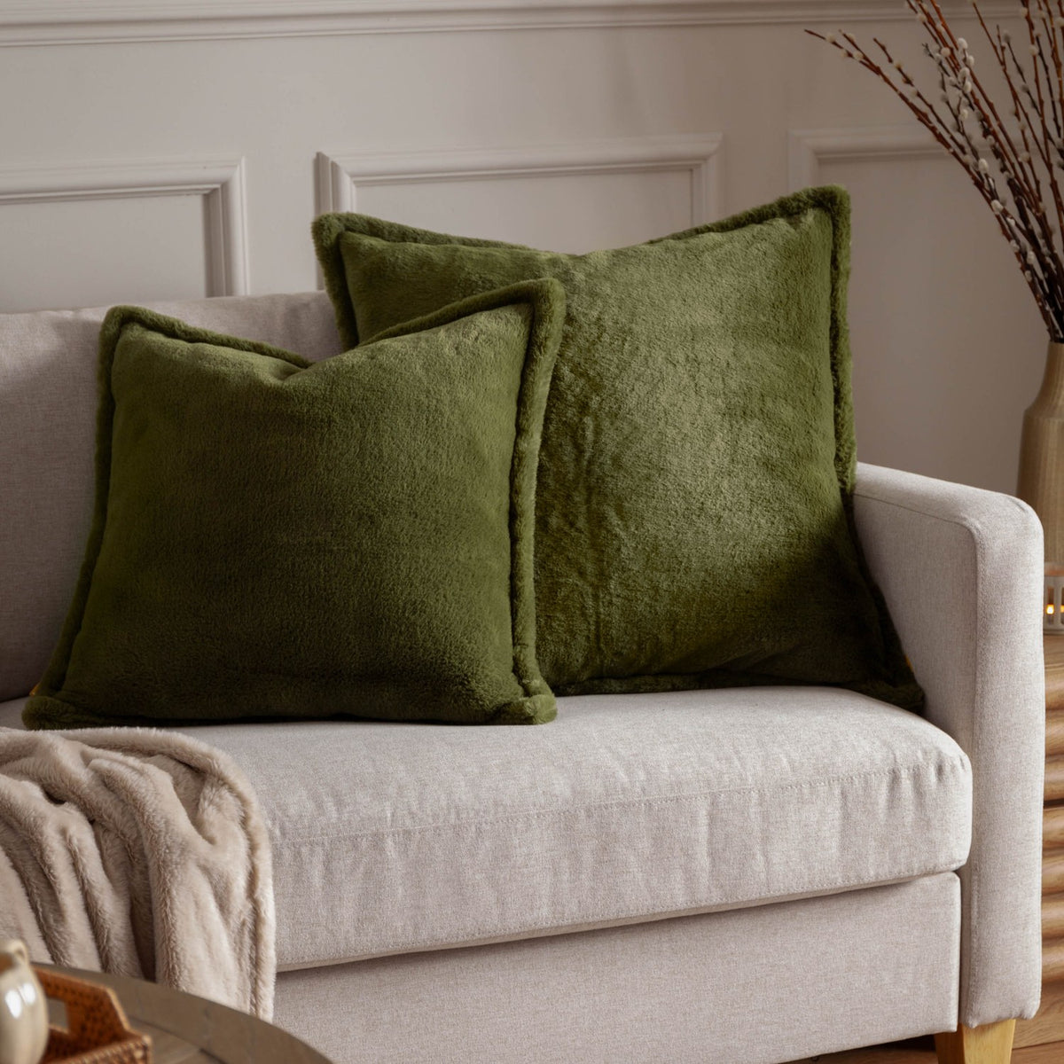 Two square Kallu Reversible Faux Fur Cushions in olive green on a light grey sofa, shown with a beige throw in a living room setting.