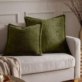 Two square Kallu Reversible Faux Fur Cushions in olive green on a light grey sofa, shown with a beige throw in a living room setting.