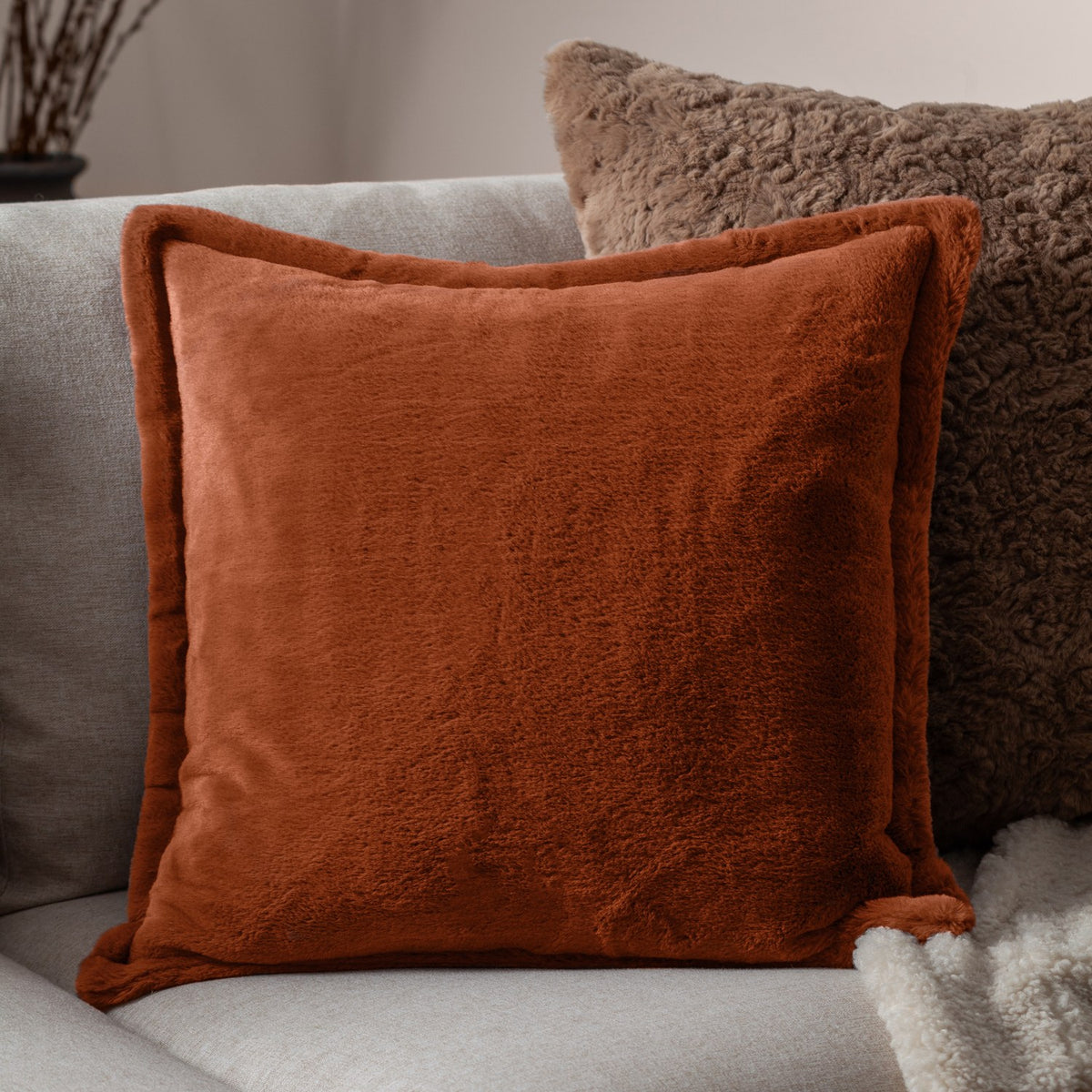 Kallu Reversible Faux Fur Cushion in rust orange with plush pile on both sides, shown on a light grey sofa beside a textured brown cushion.