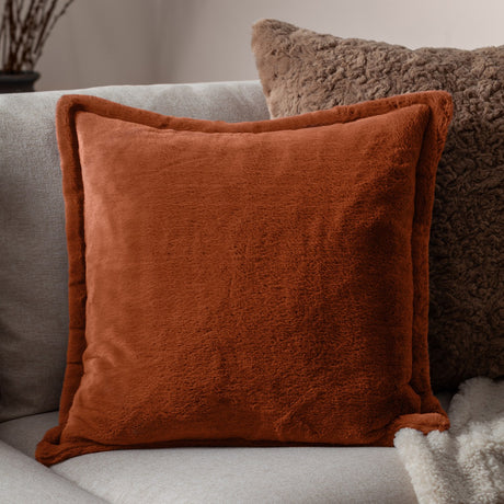 Kallu Reversible Faux Fur Cushion in rust orange with plush pile on both sides, shown on a light grey sofa beside a textured brown cushion.