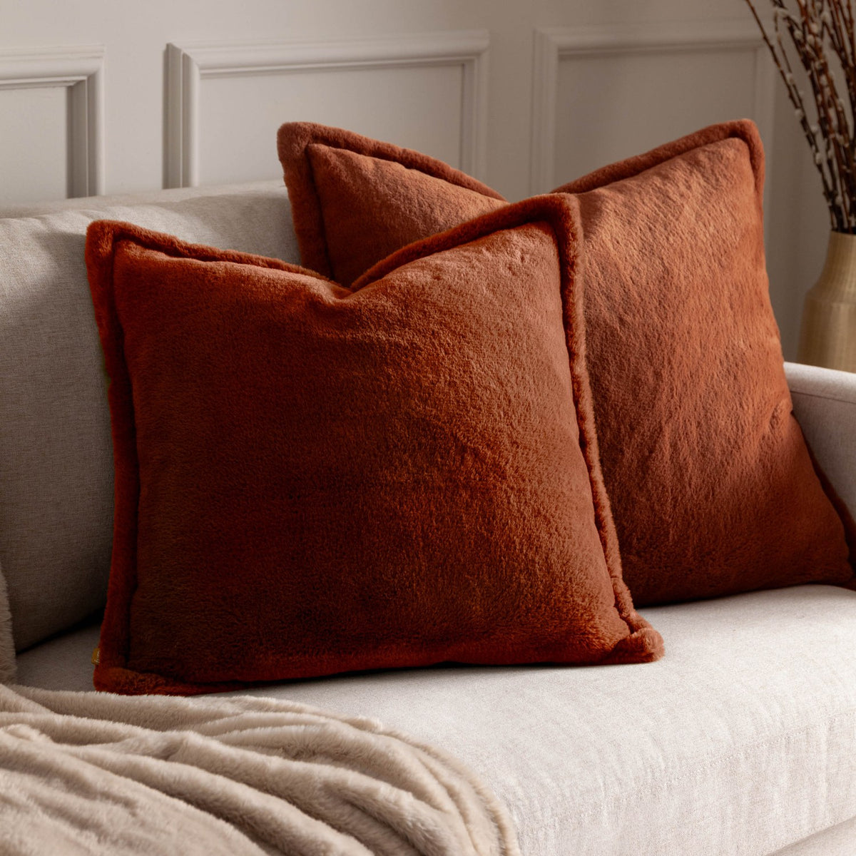 Two square Kallu reversible faux fur cushions in rust orange with a dense plush pile, shown on a light beige sofa in a living room setting.