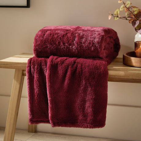 Kallu Polyester Throw in berry-red faux fur, folded with a plush pile, draped over a wooden bench in a living room setting.