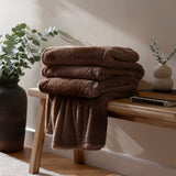 Kallu Plain Faux Fur Throw in chocolate brown, folded in layers on a wooden bench beside books and a vase, with one end draped down to show the deep pile texture.