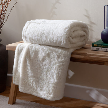Kallu Reversible Faux Fur Throw in ecru, rolled with one end draped, showing its soft cream pile, styled on a wooden bench beside books and a vase.