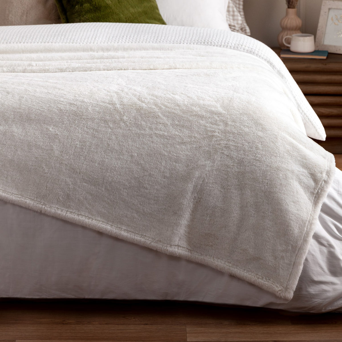 Kallu Reversible Faux Fur Throw in Ecru draped over a bedroom bed, showing the soft cream pile fabric folded to reveal its double-sided finish.