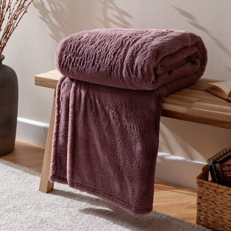 Kallu Reversible Faux Fur Throw in mulberry, folded on a wooden bench with one end draped down, showing its dense double-sided pile in a living room setting.