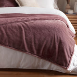 Kallu Reversible Faux Fur Throw in mulberry, dense plush pile draped over a white bed with folded edge, styled with white bedding and a wooden headboard.