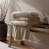 Kallu Reversible Faux Fur Throw in natural, folded on a wooden bench beside a dark vase and dried stems in a living room setting.