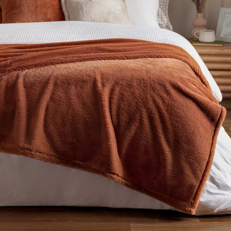 Kallu Reversible Faux Fur Throw in rust draped over a bed, showing its dense soft pile and plain woven border against white bedding.