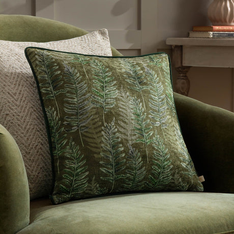 Kielder Forest Woodland Velvet Cushion in khaki with hand-painted fern print in shades of green, shown on a green upholstered armchair beside a neutral cushion.