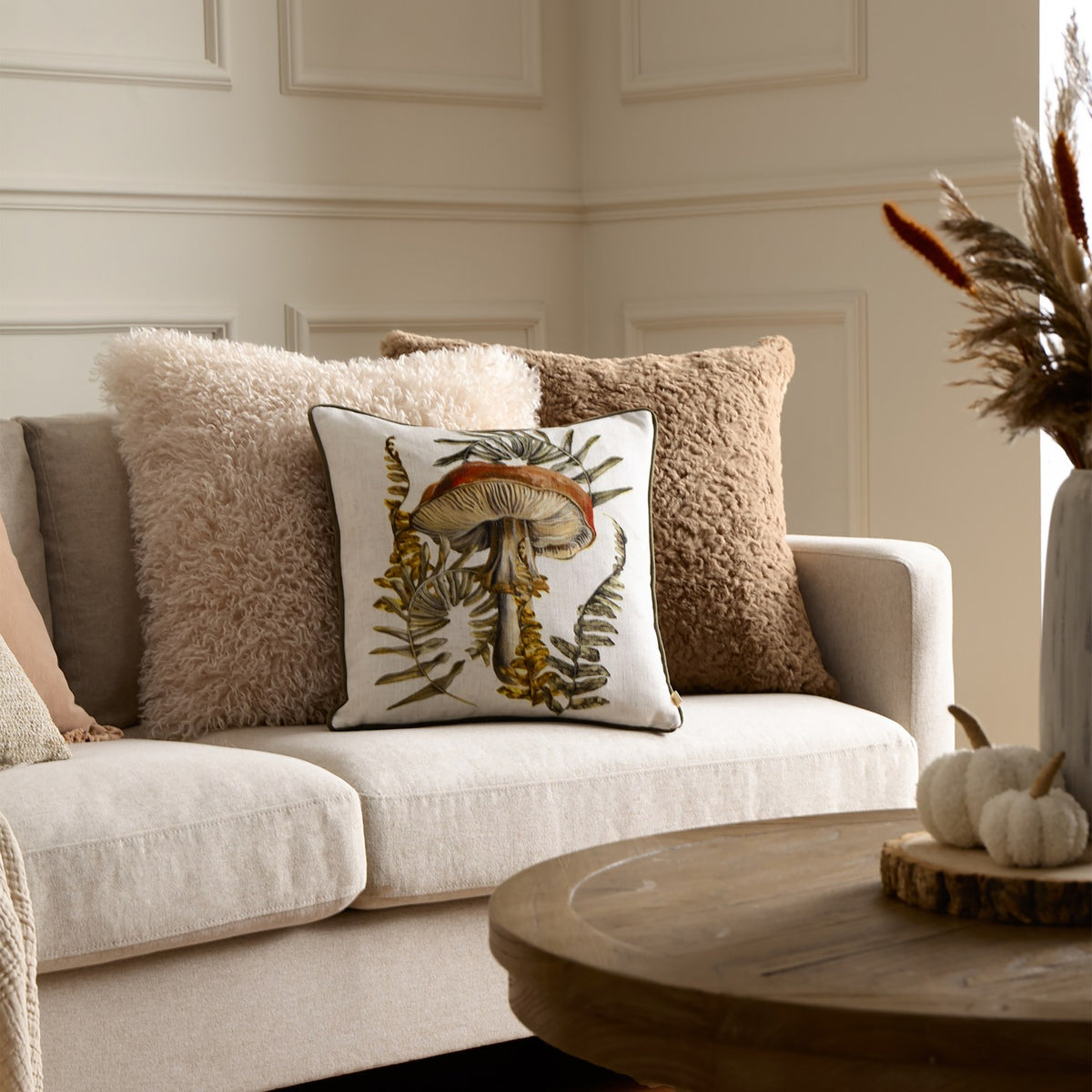 Kielder Forest Mushroom Velvet Cushion in Natural on a beige sofa, cream square cushion with a rust-cap mushroom and olive fern print, styled with fluffy textured cushions.