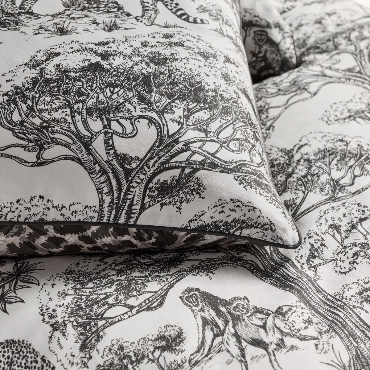 Kisumu Cotton Jungle Duvet Cover Set in monochrome, showing a pillowcase with hand-drawn Baobab tree and monkey toile print, edged with leopard-print piping.