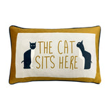 Ochre velvet lumbar cushion with cream centre panel, navy piping, and two black-and-white cats flanking The Cat Sits Here text, shown on a white background.