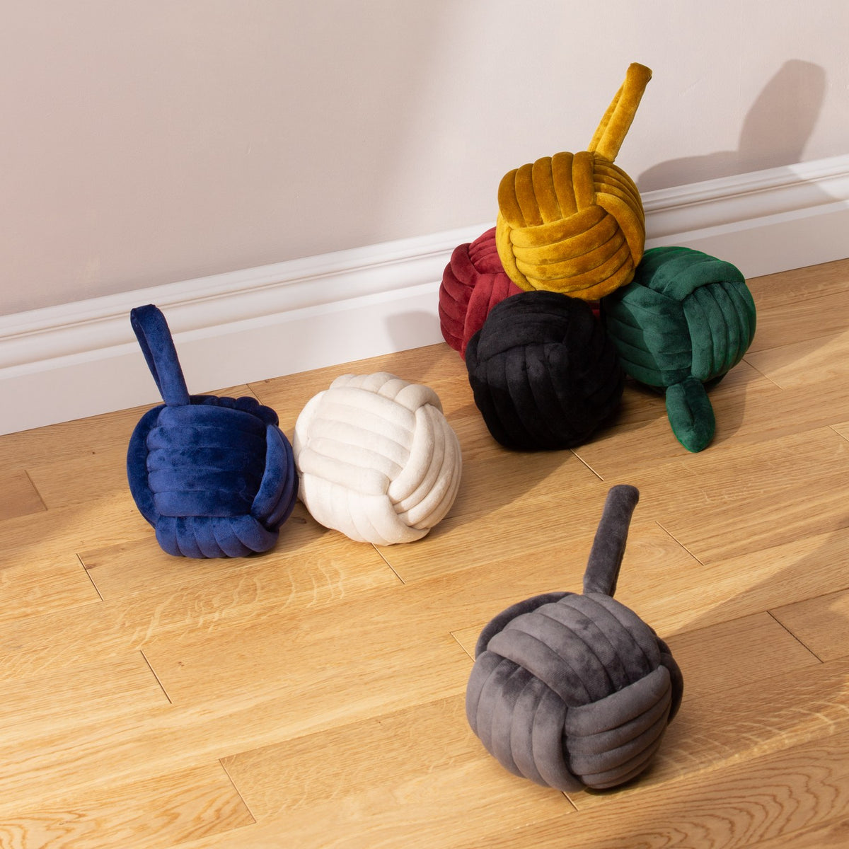 Knot Plain Velvet Door Stop in natural off-white, shown among coloured velvet knot door stops on a wooden floor, with an interlaced ball design and looped top handle.