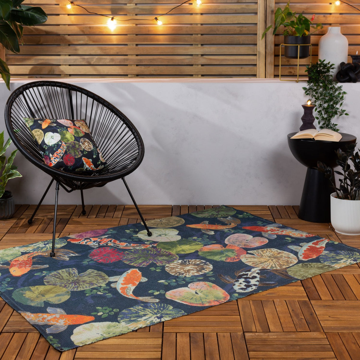 Koi Pond Animal Polyester Rug in midnight blue with orange and white koi carp and lily pad print, laid on a wooden deck beside a black rattan chair.