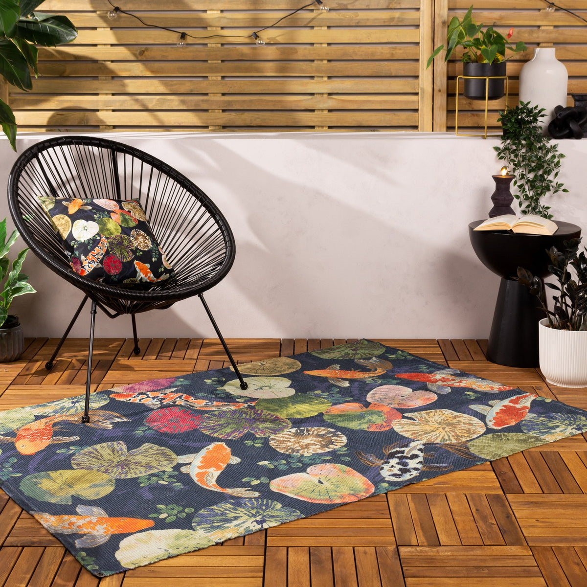 Koi Pond Animal Polyester Rug in Midnight, a navy flatweave rug with orange and white koi carp and lily pads, shown on a wooden deck beside a black woven chair.