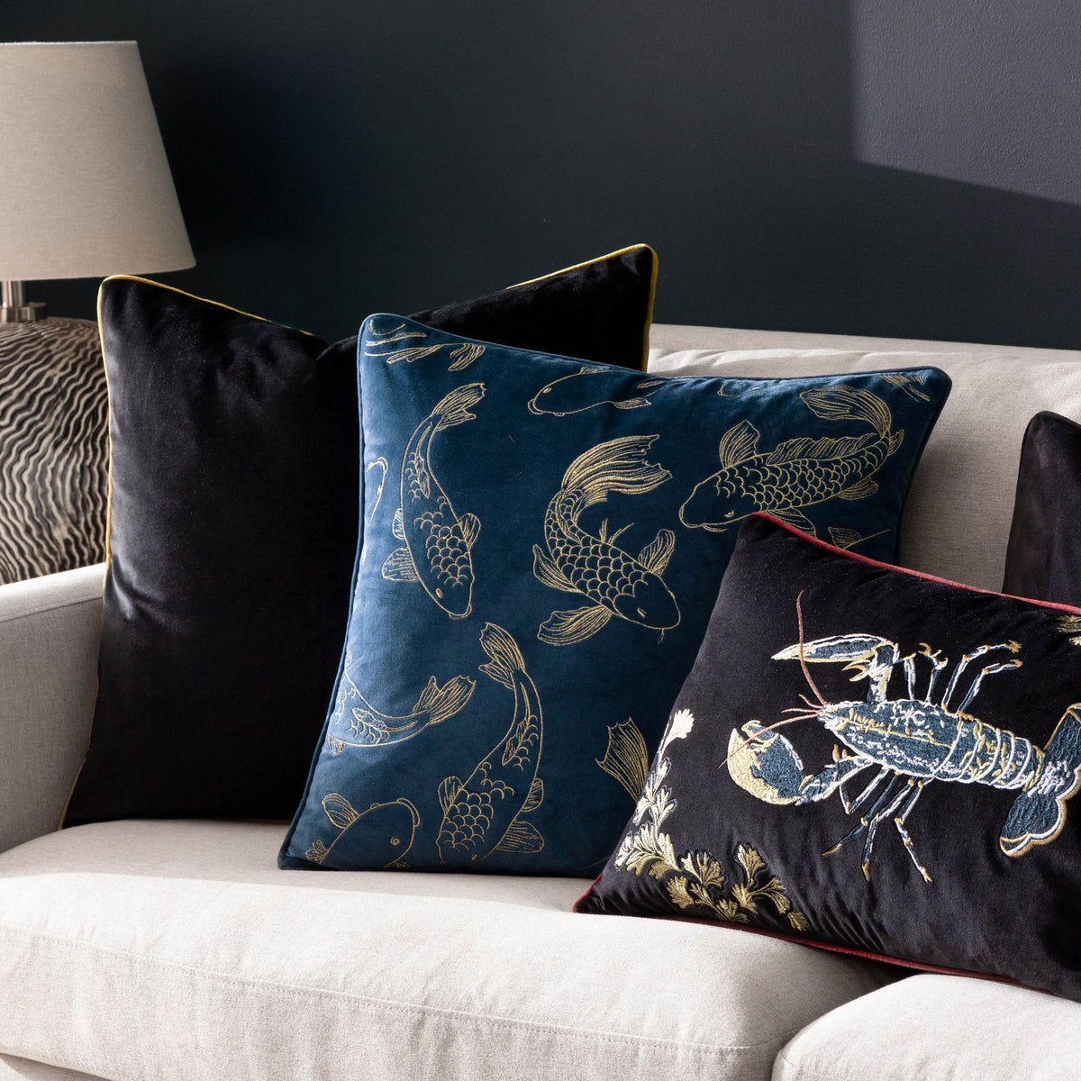 Navy velvet Koi Pond cushion embroidered with gold koi carp, shown on a sofa alongside black velvet and lobster-embroidered cushions.