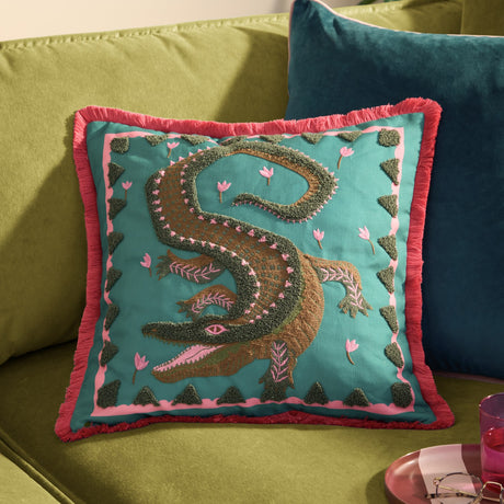Teal polyester cushion with tufted crocodile design in green and brown, pink floral accents, pink inner border and coral fringe trim, shown on a green sofa.