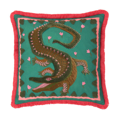 Square teal cushion with tufted crocodile design in green and brown, pink flower accents, pink inner border and coral fringe trim, shown on a white background.