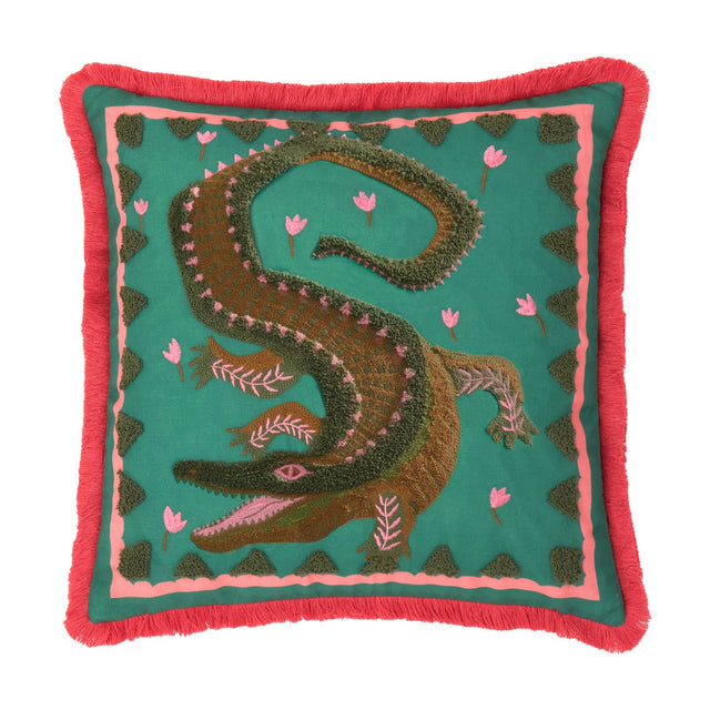 Square teal cushion with tufted crocodile design in green and brown, pink flower accents, pink inner border and coral fringe trim, shown on a white background.