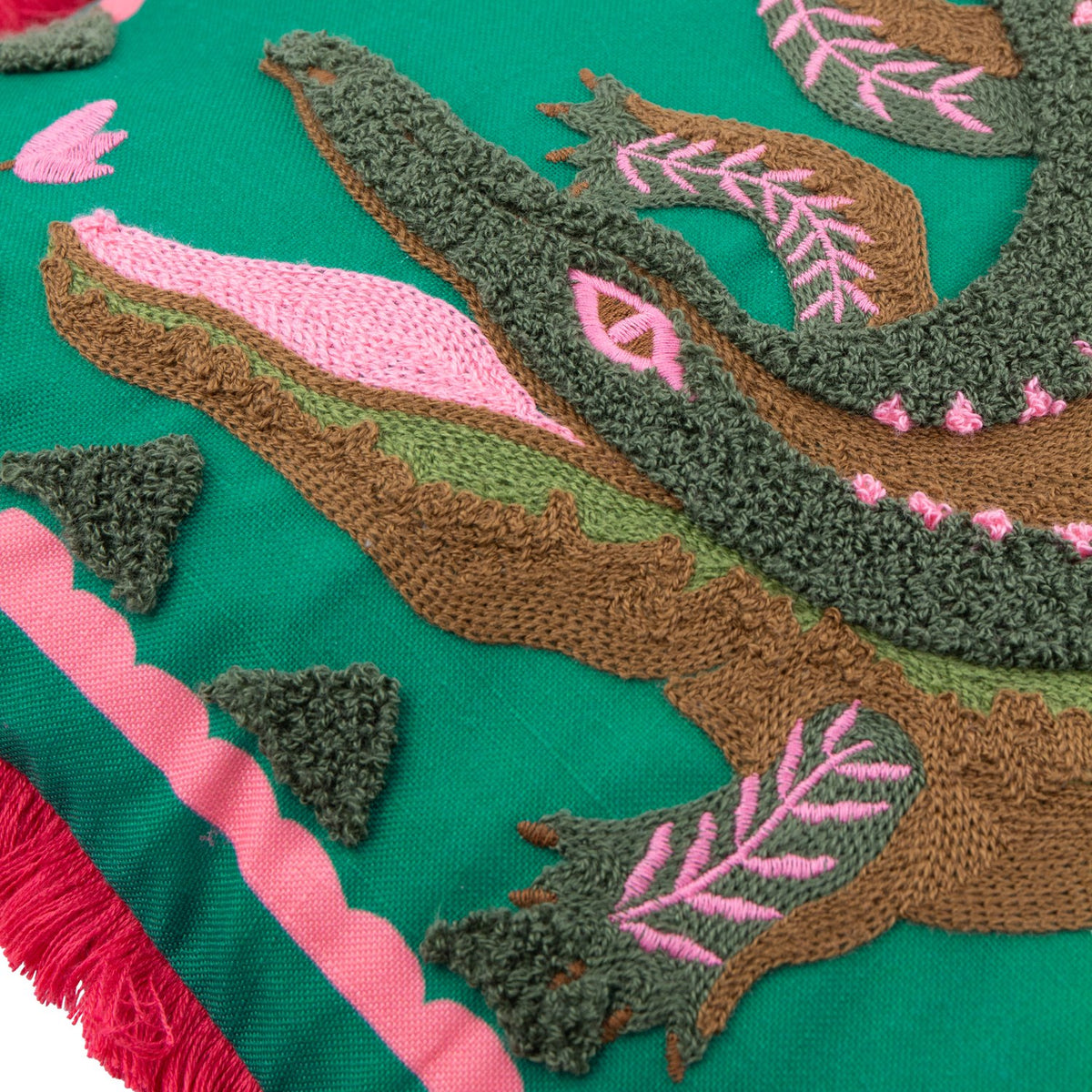 Close-up of the Les Animaux Croc Monsieur cushion showing tufted crocodile embroidery in green and brown with pink eye and leaf details on a teal ground, edged with a pink border and coral fringe.