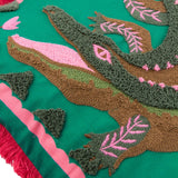 Close-up of the Les Animaux Croc Monsieur cushion showing tufted crocodile embroidery in green and brown with pink eye and leaf details on a teal ground, edged with a pink border and coral fringe.