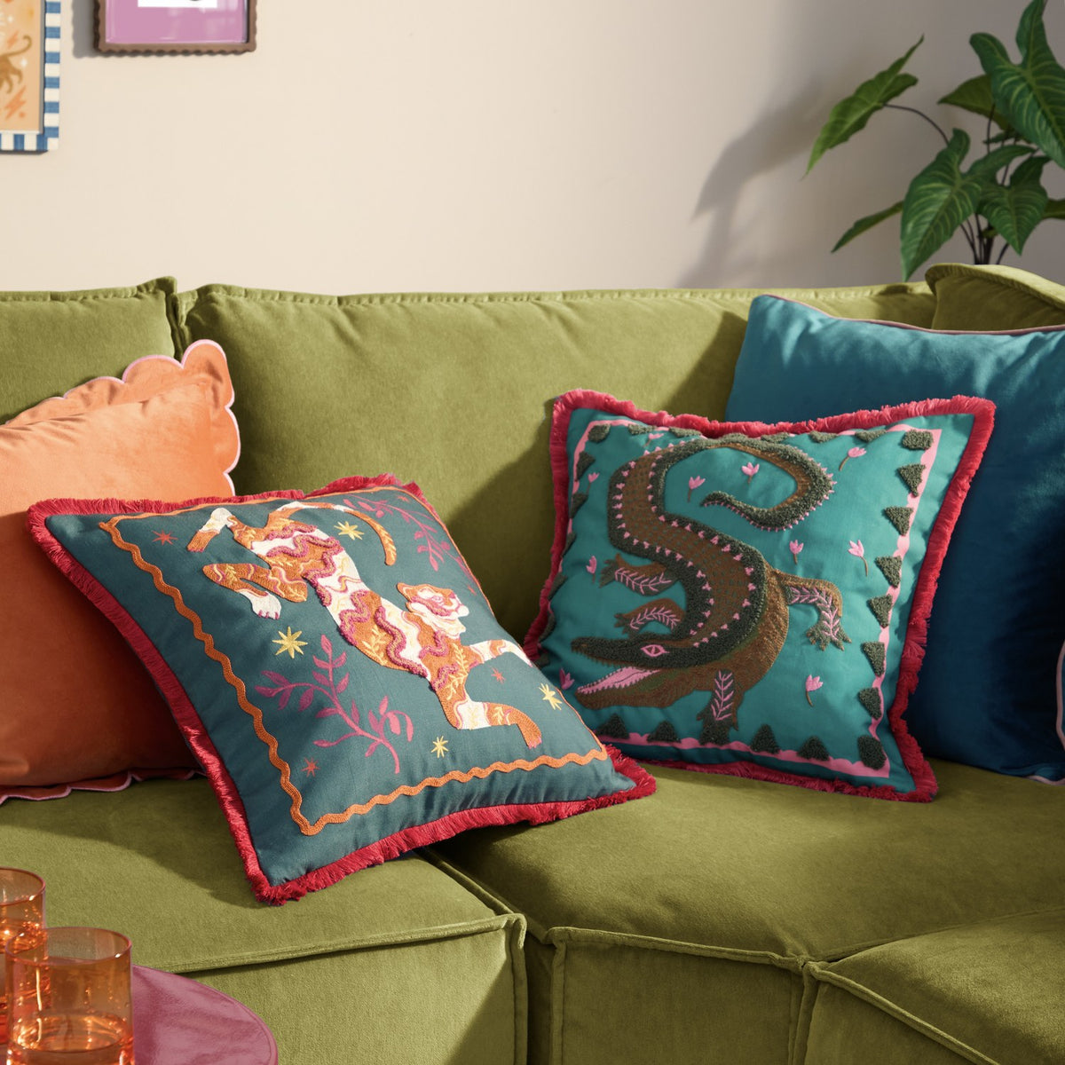 Teal polyester cushion with tufted crocodile design in green and brown, pink floral accents, pink inner border and red fringe trim, shown on a green sofa beside a tiger-print cushion.