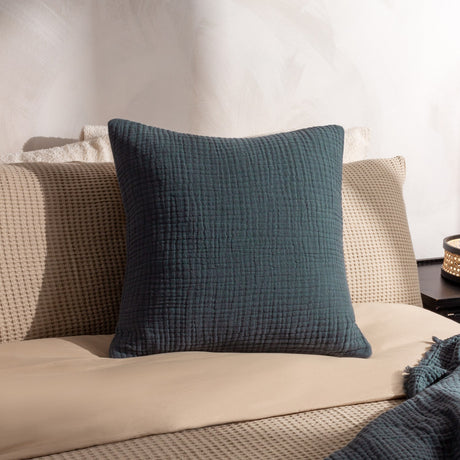 Lark Reversible Cotton Cushion in Dusk teal-blue with crinkled muslin texture, styled on a beige waffle-weave sofa against a light neutral wall.