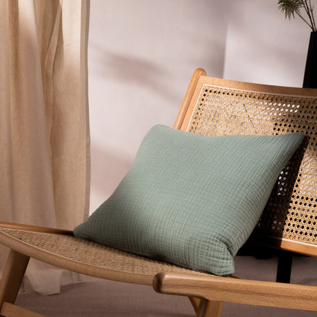 Lark Reversible Cotton Cushion in eucalyptus green, crinkle-textured muslin cotton, 45 x 45cm, shown on a rattan and wood chair.