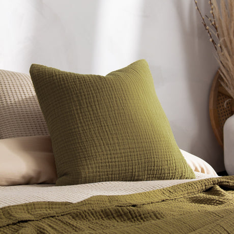 Lark Reversible Cotton Cushion in khaki green, 45 x 45cm square with textured muslin crinkle cotton surface, shown propped against pillows on a bedroom bed with a matching woven throw.