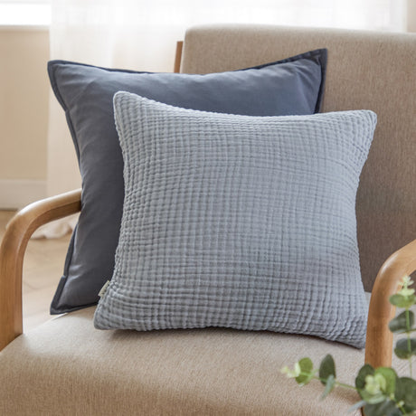 Lark Plain Cotton Cushion in mineral blue with crinkled muslin texture, shown 45 x 45cm on an armchair alongside a slate blue velvet cushion.