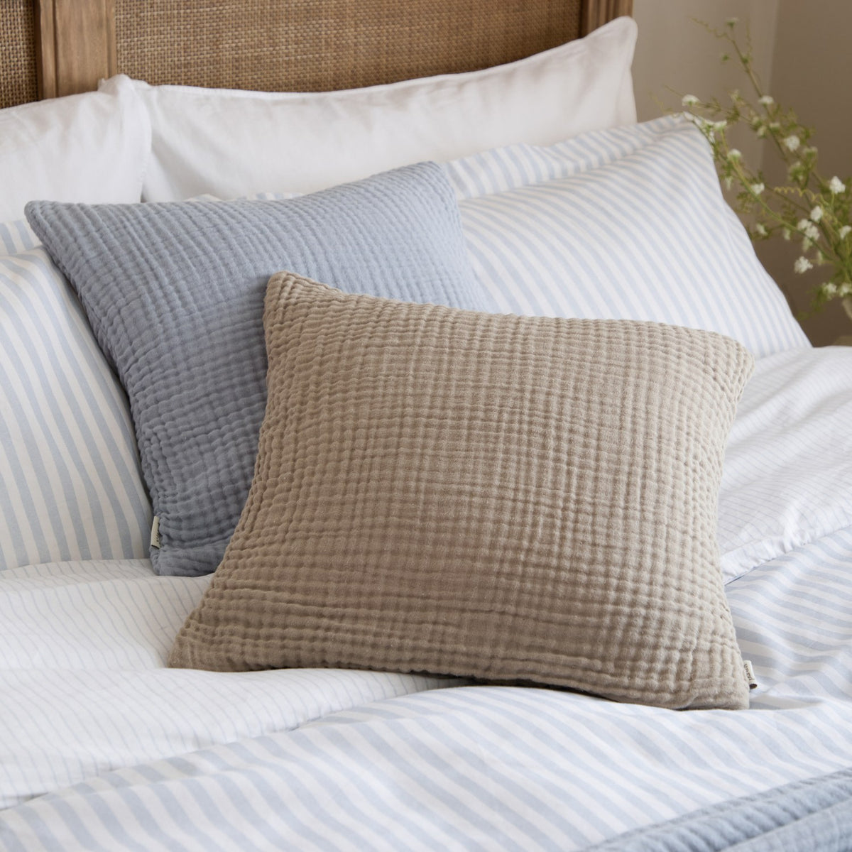 Lark Plain Cotton Cushion in Mineral Blue, a square 45 x 45cm crinkled muslin cushion with a textured ribbed surface, shown on a bed layered with a taupe cushion and striped bedding.