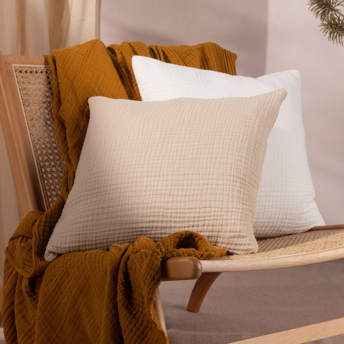 Lark Reversible Cotton Cushion in natural beige crinkled muslin, shown with a white cushion and mustard throw on a rattan chair.