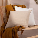 Lark Reversible Cotton Cushion in natural beige crinkled muslin, shown with a white cushion and mustard throw on a rattan chair.