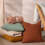 Terracotta and sage muslin cotton cushions styled with a folded natural cotton throw and rolled pink blanket beside a rattan chair, showing the Lark cushion's crinkled four-layer texture.