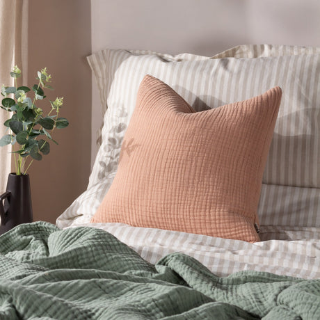 Lark Reversible Cotton Cushion in Pink Clay, crinkled muslin-textured square cushion styled on a bed with striped bedding and a green throw.