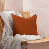 Lark Reversible Cotton Cushion in pecan rust, crinkled muslin texture, styled on a cane and wood chair with a white throw against a light background.