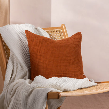 Lark Reversible Cotton Cushion in pecan rust, crinkled muslin texture, styled on a cane and wood chair with a white throw against a light background.