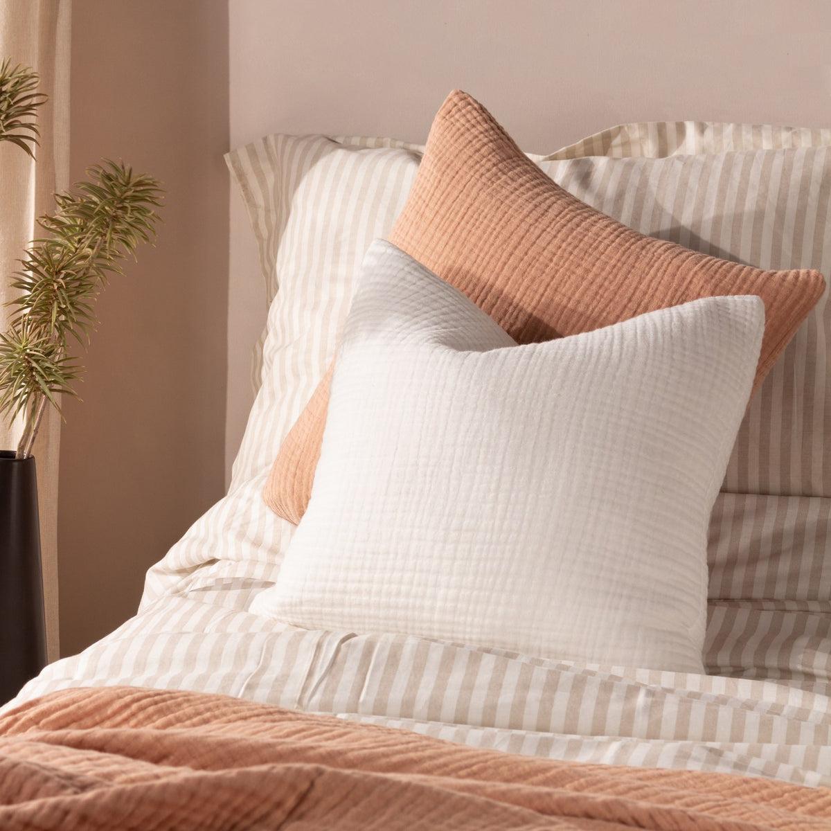 Lark Reversible Cotton Cushion in white, textured muslin crinkle cotton, 45 x 45cm square, styled at the front of a bed with striped bedding and a terracotta cushion behind.
