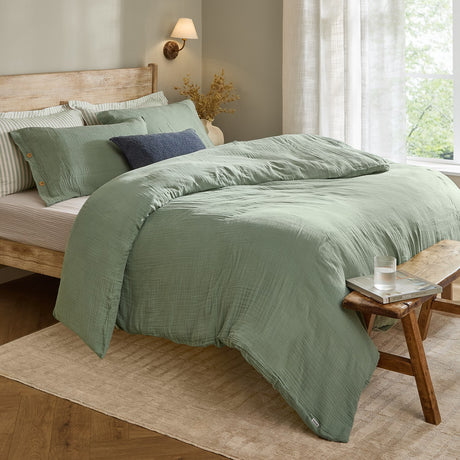 Lark Cotton Plain Duvet Cover Set in eucalyptus green, crinkled muslin cotton bedding with wooden button fastenings, styled on a wooden bed in a bedroom setting.