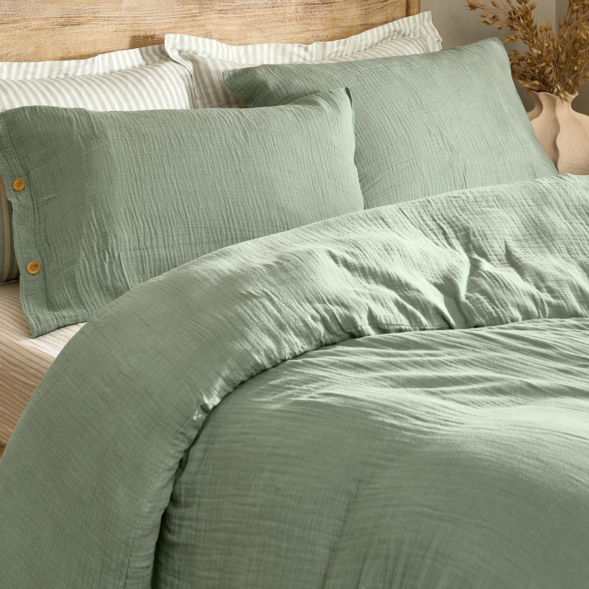 Lark Cotton Plain Duvet Cover Set in eucalyptus green, crinkled muslin cotton with wooden button fastening, styled on a bed with striped pillowcases in a bedroom setting.